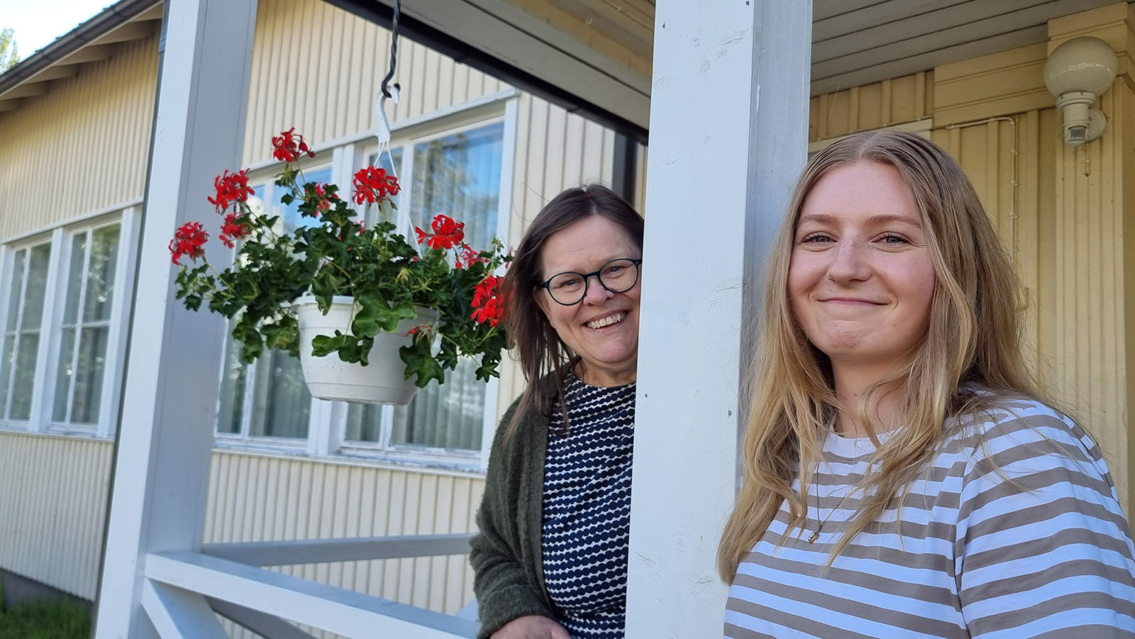 Kaksi naista hymyilee kameralle ulkona pylvään molemmin puolin.